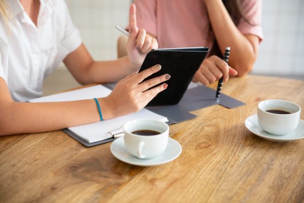 Business ladies meeting at table, watching presentation on tablet, discussing project or deal. Cropped shot of table and hands. Consulting or cooperation concept